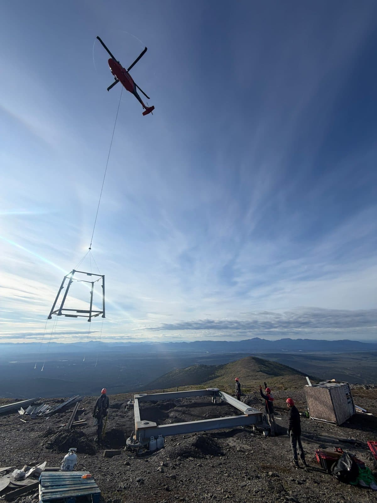 Helicopter long-lining a tower section to a hilltop foundation while the ground crew in hard hats guides the lift.