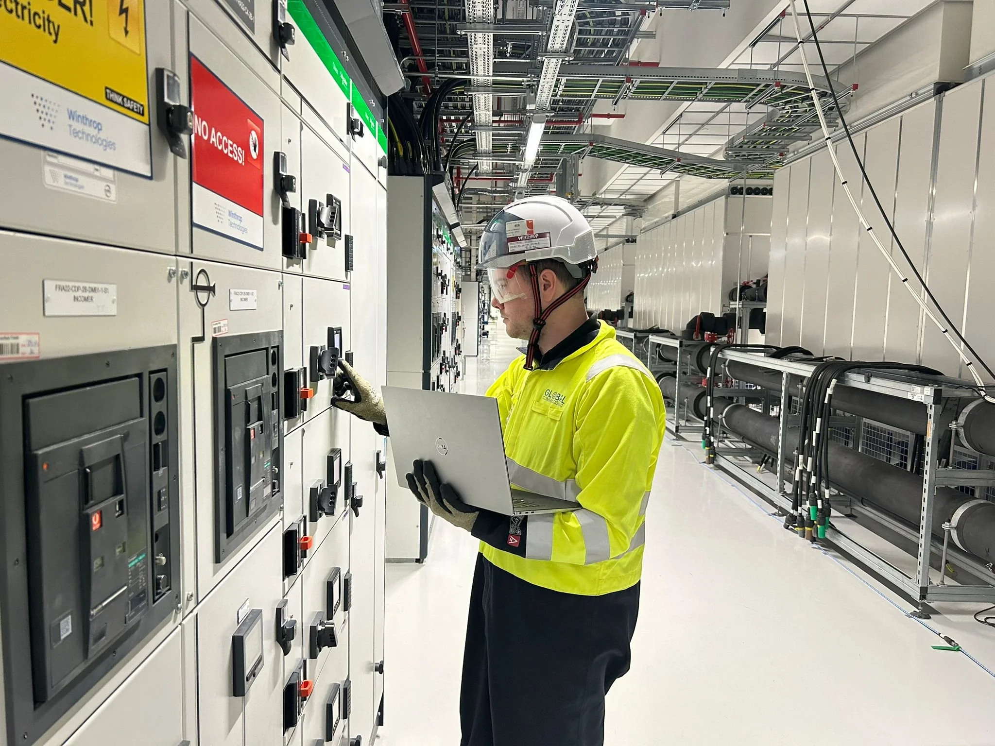 Commissioning technician with laptop checking electrical switchgear panels in a live data hall.