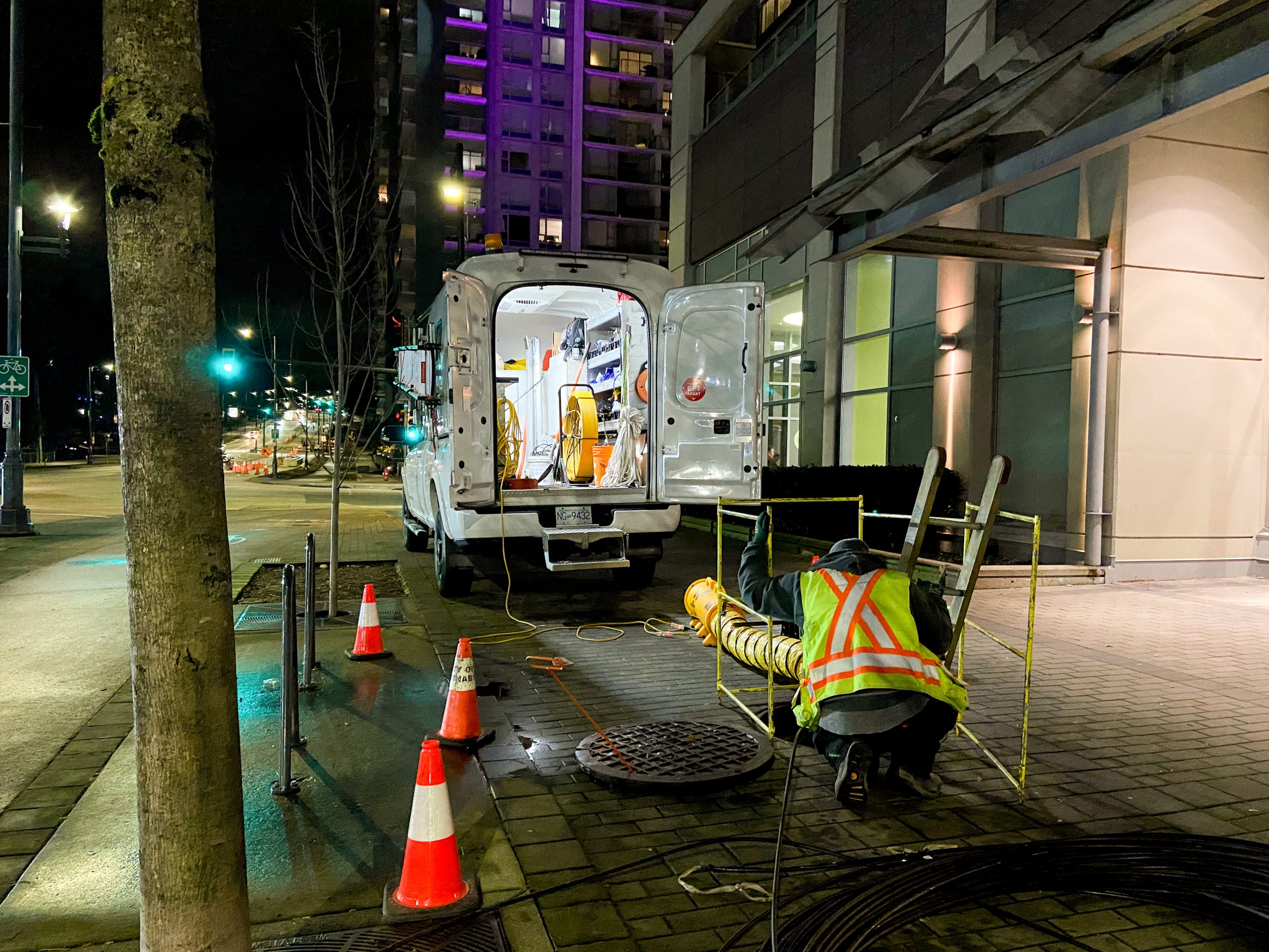 Night fiber splicing operation at a manhole with work van and traffic cones in an urban setting.